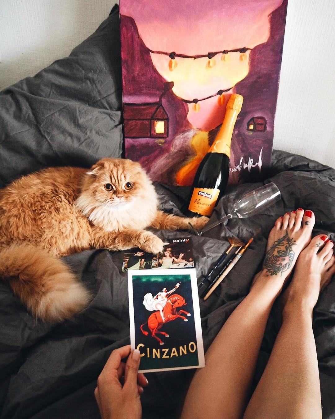 Фото Девушка сидит на постели, рядом лежит рыжий кот и стоит у рисунка бутылка шампанского