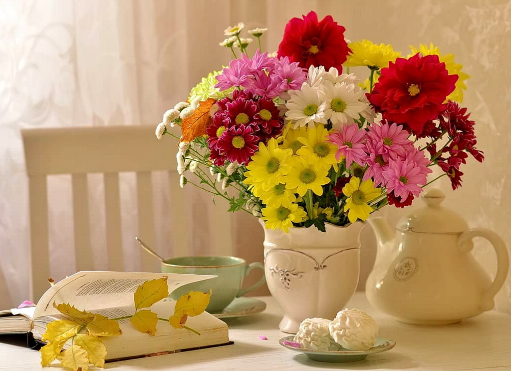 Фото Букет, составленный из разных красивых цветов, в белой фарфоровой вазе на столе рядом с открытой книгой, на страницах которой лежит осенняя веточка, зефиром на блюдечке и фарфоровой посудой