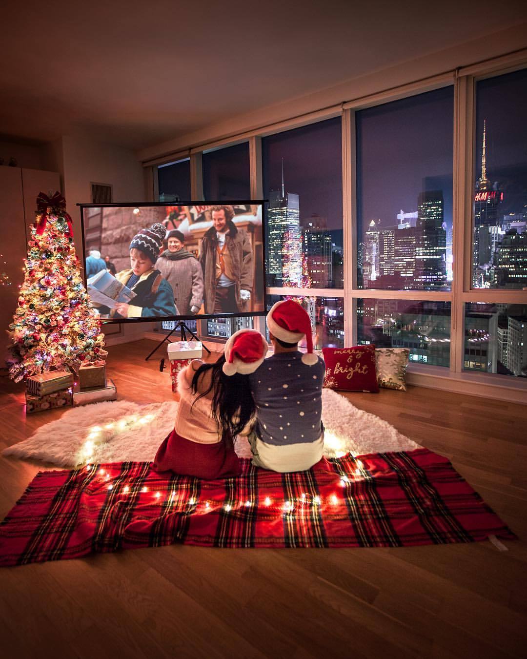Фото Пара в новогодних шапочках, постелив плед посреди комнаты, смотрит фильм Один дома, рядом стоит украшенная елка с подарками