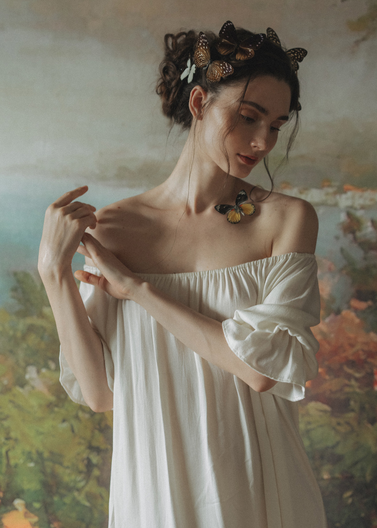 Фото Модель Олеся Петрова в белом платье с бабочками на волосах и у шеи стоит на фоне картины