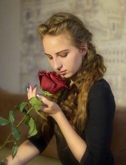Фото Девушка в черном платье стоя в аудитории на фоне стены нюхает красную розу держа ее в руке