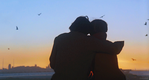 Анимация Парень и девушка сидят обнявшись на берегу моря, вокруг них летают чайки, вдали видны здания, гифка Парень и девушка сидят обнявшись на берегу моря, вокруг них летают чайки, вдали видны здания
