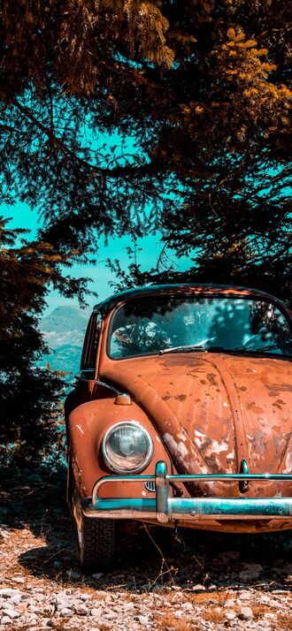 обои на телефон Ржавый коричневый автомобиль Volkswagen Beetle стоит на каменистой земле на фоне темных деревьев, яркого голубого неба и далеких гор