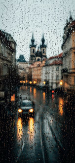 Обои на телефон Прага / Prague, Чехия / Czech Republic, капли дождя на стекле, через которое виден автомобиль с включенными фарами на мокрой дороге, на фоне размытых старинных зданий и церкви с двумя шпилями
