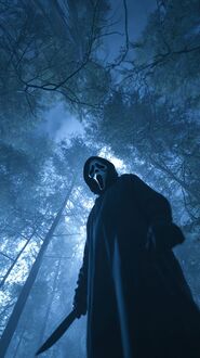 Обои на телефон Персонаж Призрачное лицо / Ghostface из фильма Крик / Scream в черном костюме и маске держит нож, на фоне темного леса с высокими деревьями, освещенного синим светом
