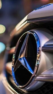 Обои на телефон Крупный план блестящего хромированного логотипа Mercedes-Benz на решетке радиатора автомобиля, на фоне размытых городских огней