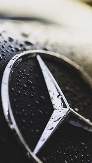 Обои на телефон Серебристый логотип Mercedes-Benz с каплями воды на черной поверхности автомобиля на фоне размытого света